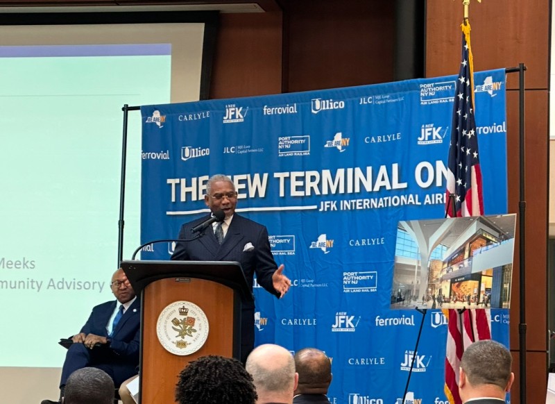 The image shows a formal event where a man in a suit is speaking at a podium. The podium has a seal with a crown and a plant below it. Behind him is a blue backdrop featuring various logos, including "Carlyle," "Ullico," and "Port Authority NY NJ." The backdrop prominently displays the phrase "THE NEW TERMINAL ONE JFK INTERNATIONAL." To the right of the speaker, an American flag stands next to a photograph or rendering of an airport terminal. A seated man in a blue suit listens attentively to the left.