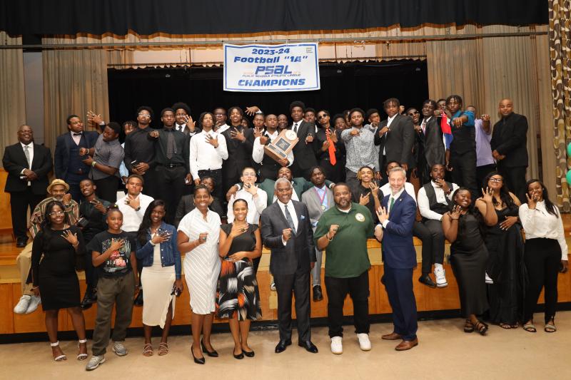 A large group of people are gathered on a wooden stage, dressed in formal and semi-formal attire. The group consists of men and women, primarily young adults, with some older individuals. They are posing for the camera, mostly smiling, and many are wearing or holding rings prominently on their fingers. In the center, an individual is holding a plaque. Above them, a banner hangs with text indicating a sports achievement.